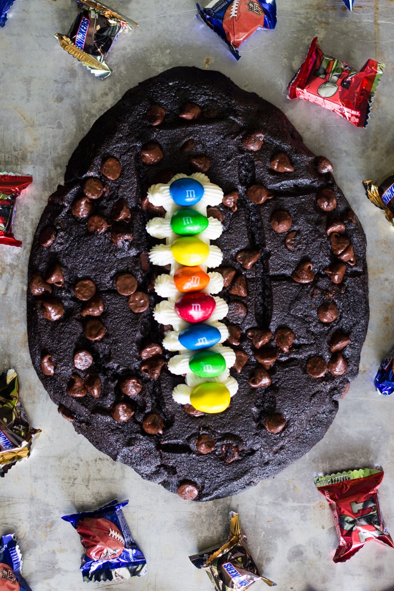 Made with chocolate chocolate chip cookie dough, vanilla frosting, and M&M's, this fun football cookie cake is very easy to make and perfect for parties.