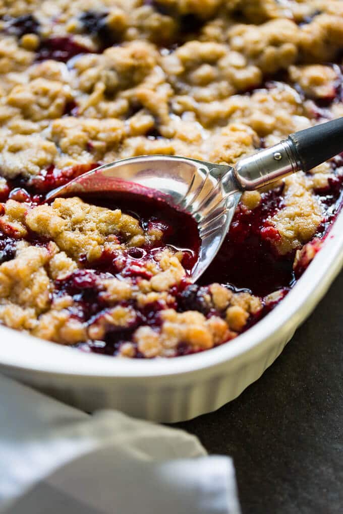 White casserole dish filled with blackberry crumble and a silver serving spoon with a wooden handle