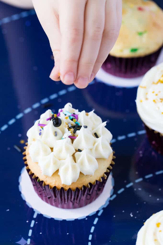 Child's fingers sprinkling decorations on a cupcake with vanilla frosting