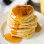Stack of small pancakes with ham chunks on a white plate with maple syrup pouring over