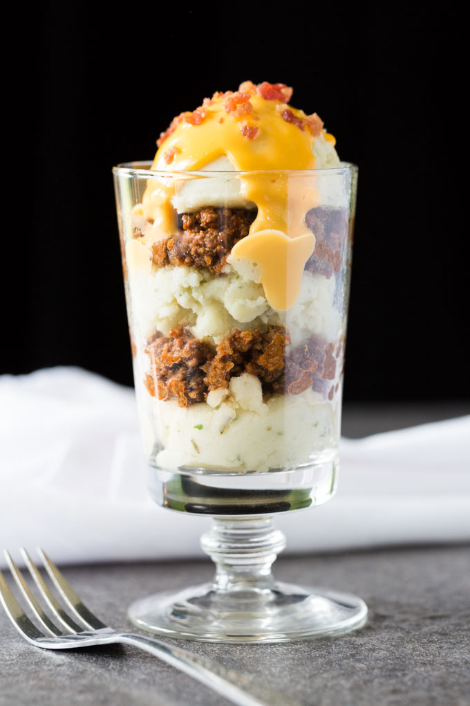 Parfait glass filled with mashed potatoes, chili, cheese sauce, and bacon