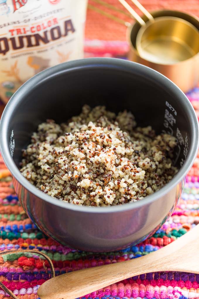 Cooked tricolor quinoa in a rice cooker pot next to measuring cups, a rice paddle, and a bag of quinoa