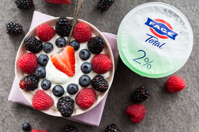 Pink bowl of Greek yogurt topped with strawberries, blackberries, blueberries, and raspberries next to FAGE yogurt cup