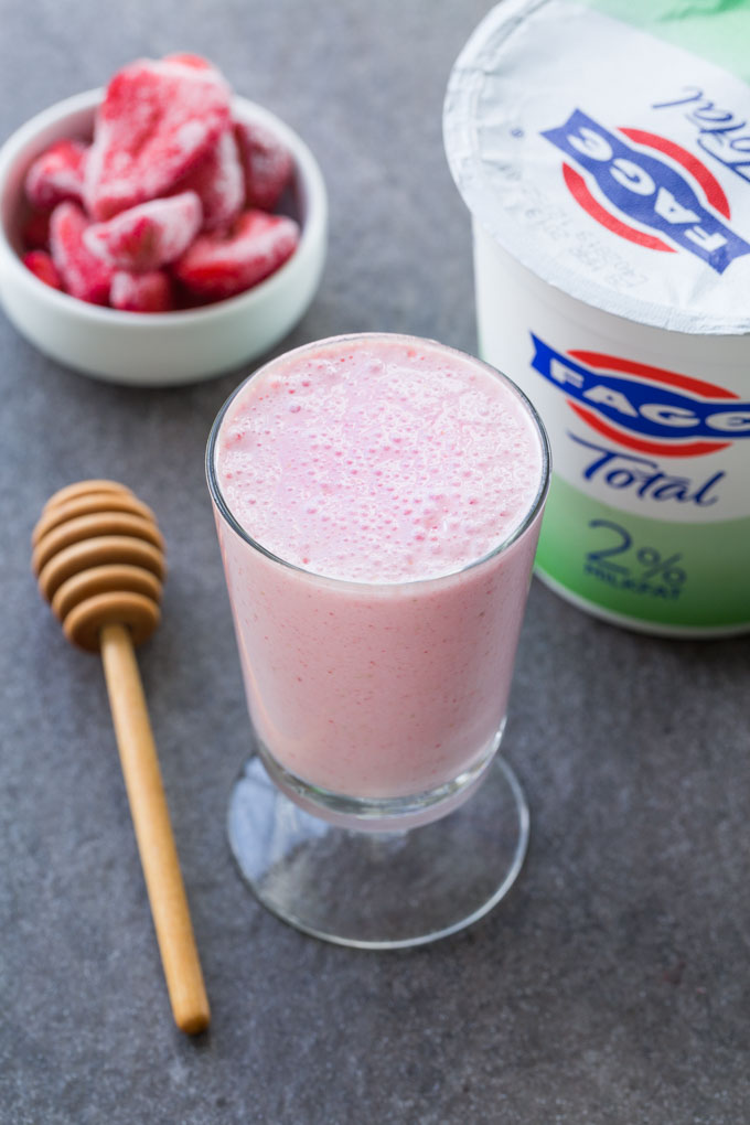 Strawberry lassi in a footed glass, FAGE Total Greek Yogurt, frozen strawberries in a white bowl, and a wooden honey dipper