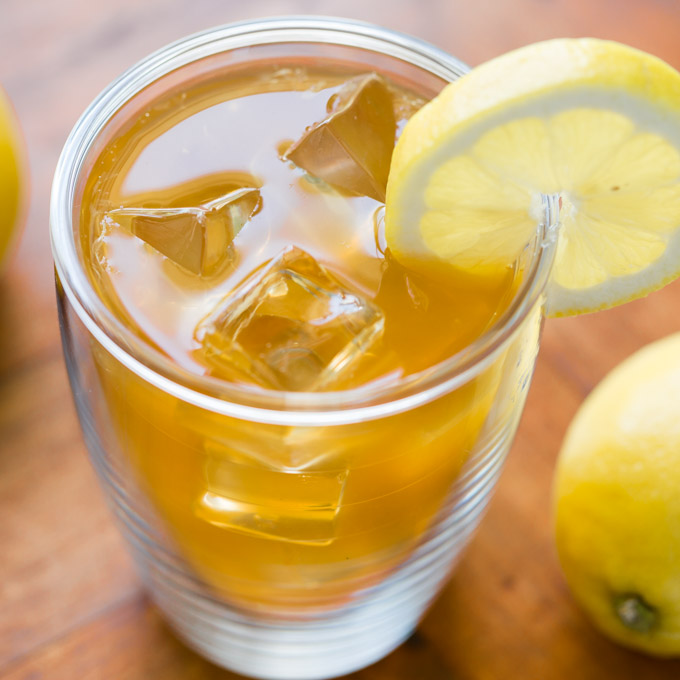Clear insulated mug of lemonade iced tea with lemon slice next to a whole lemon on a wood surface