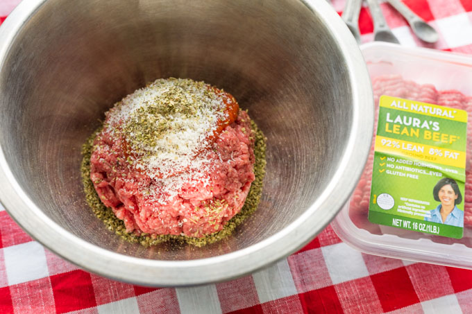 Mixing bowl with ground beef, parmesan, tomato sauce, and Italian seasoning on a red checked tablecloth with package of Laura's Lean Beef