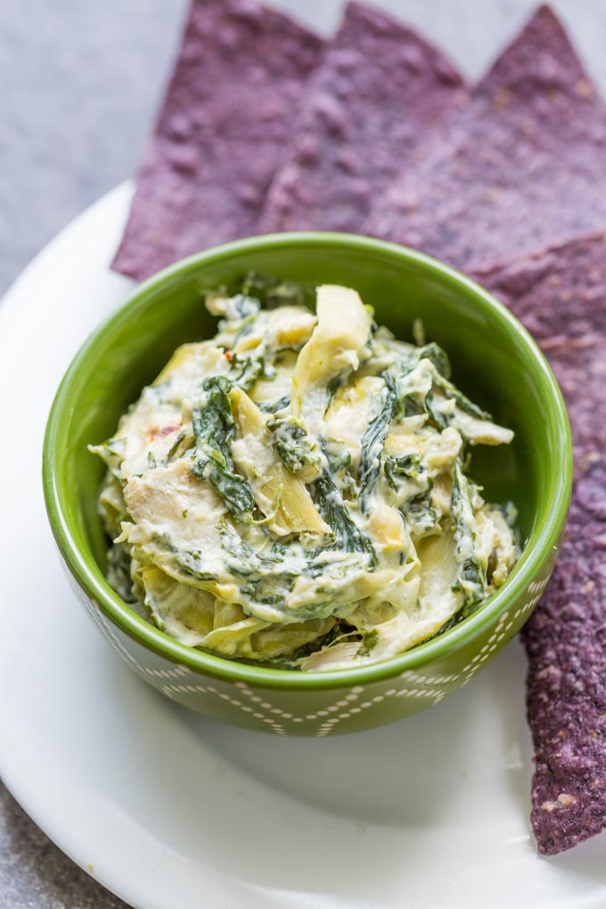 Hot spinach artichoke dip in a green bowl on a white plate with blue corn tortilla chips